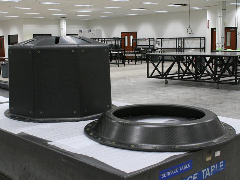 Image shows the two pieces of the smallsat launch adapter structures in a manufacturing bay. They are sitting on a granite table with a protective white foam layer on its surface. The structural ring is in the foreground and the octagonal structure is behind and to the left of the ring.